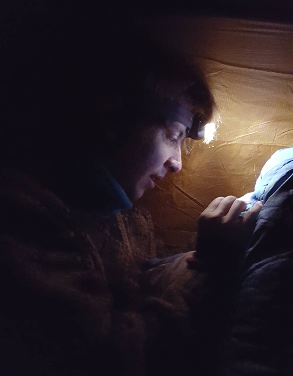 A picture of myself fiddling with a sleeping bag in a dark tent, illuminated only by me headlamp.